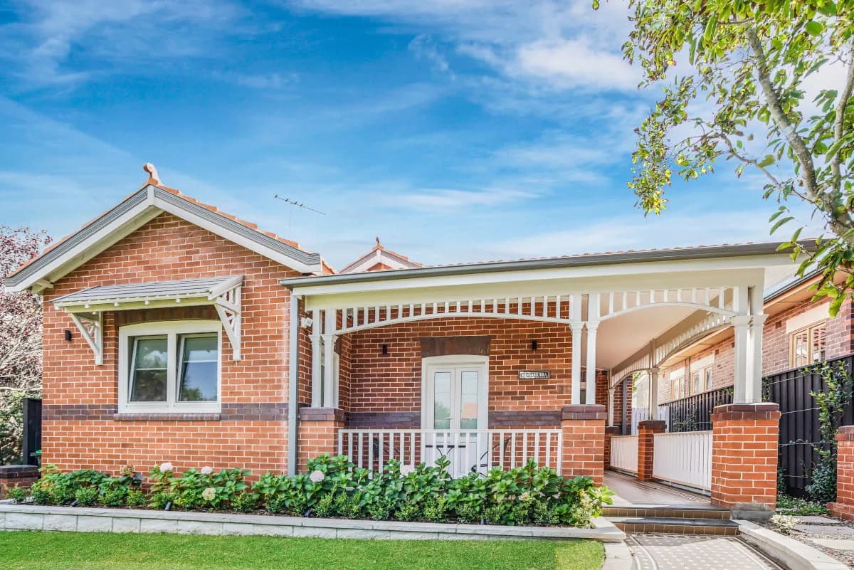Street frontage of a Federation-era red-brick bungalow with white-painted bullnose verandah, timber fretwork and established front garden