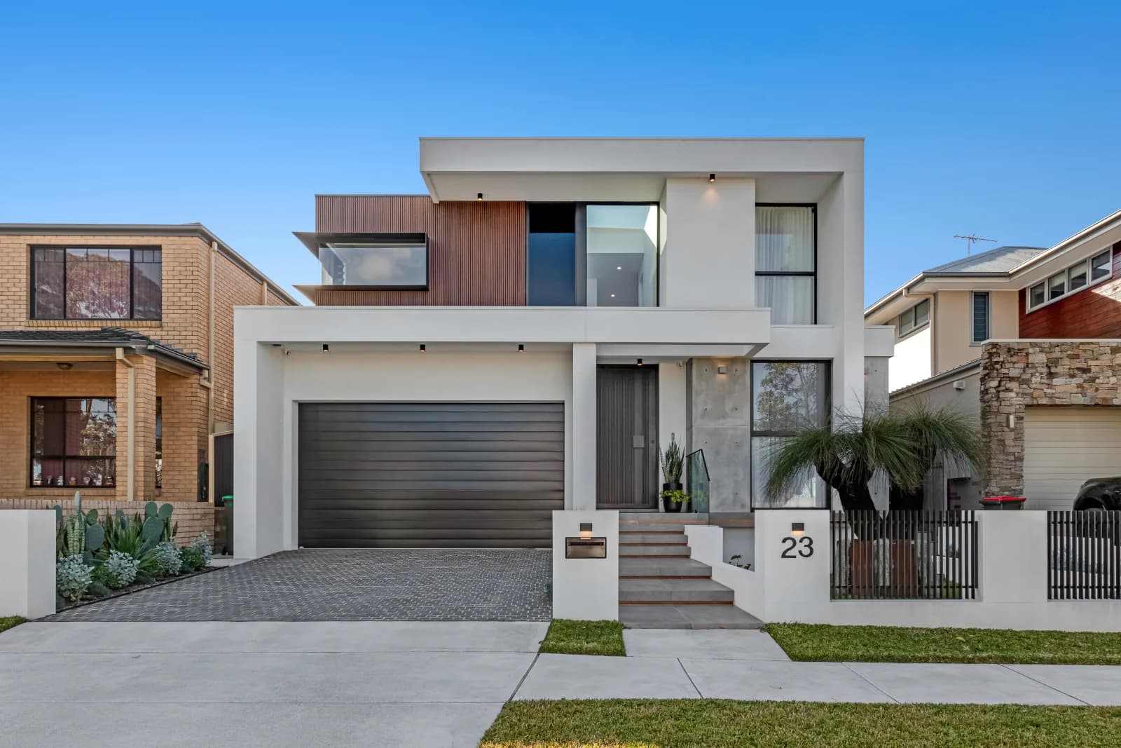 Street-facing exterior of Concord Manor new build showing a two-storey rendered façade with warm timber cladding feature panel, dark garage roller door and black iron entry gate